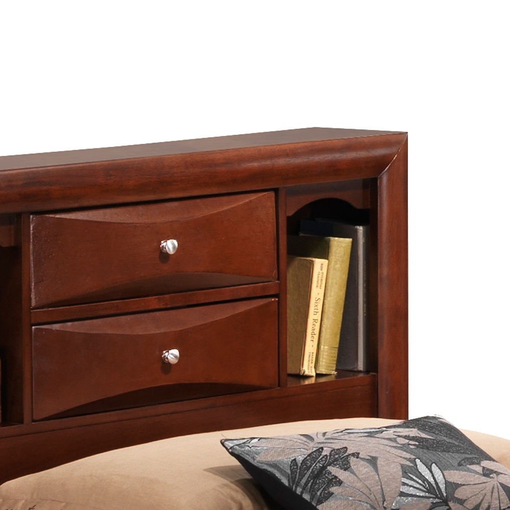 A close-up of the Marilla Storage Bed's wooden bookcase headboard shows two drawers with silver knobs and an open shelf with books, while a beige pillow and a black-and-grey cushion are in the foreground.