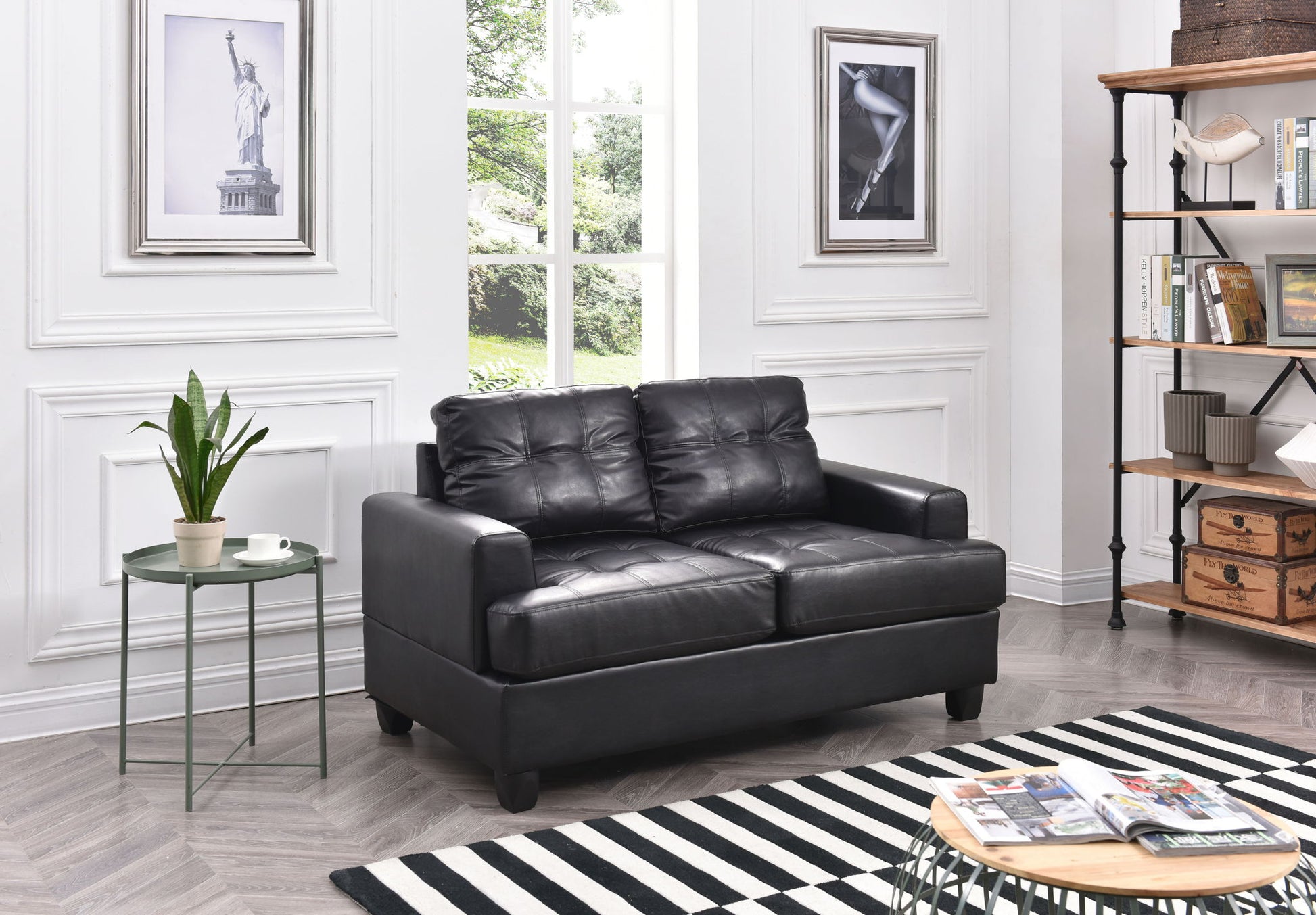 A modern living room features the Stylish Transitional Loveseat For Two in black leather, a small round green table with a potted plant and mug, striped rug, art on white-paneled walls, plus a large window and bookshelf in the background.