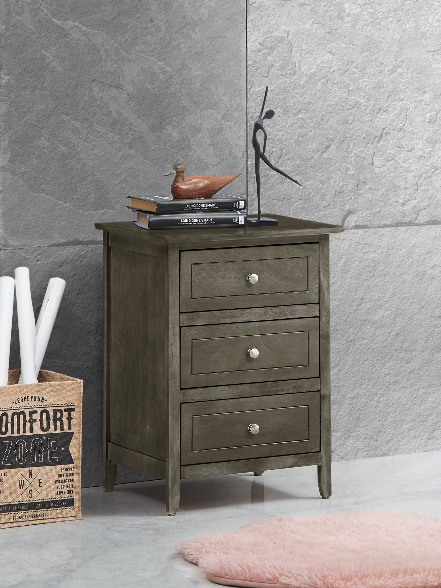 The Daniel - 3 Drawer Nightstand, crafted in gray wood with silver knobs, stands on a marble floor. On top are books, a duck figurine, and a black sculpture. Nearby: rolled papers, box, pink rug. Gray stone wall behind.