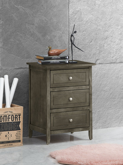 The Daniel - 3 Drawer Nightstand, crafted in gray wood with silver knobs, stands on a marble floor. On top are books, a duck figurine, and a black sculpture. Nearby: rolled papers, box, pink rug. Gray stone wall behind.