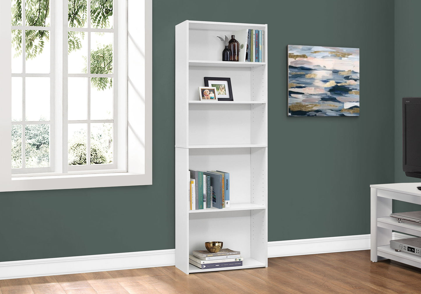 A 6-tier white bookshelf stands by a green wall with wood floors near a window, displaying books, decor, and a photo. A painting hangs nearby.