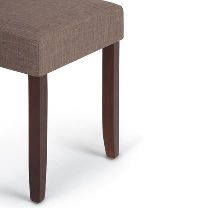 A close-up of the Acadian - Upholstered Parson Dining Chair (Set of 2) shows its brown upholstered seat and three dark wooden legs against a white background, highlighting only the seat and part of the legs.