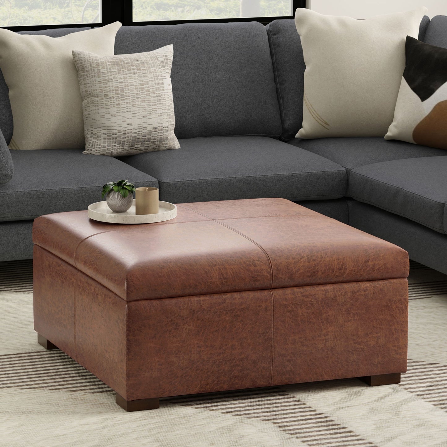The Gabbie Coffee Table Upholstered Storage Ottoman, topped with a tray holding a small plant and books, sits before a dark gray sectional with assorted pillows on a light patterned rug.
