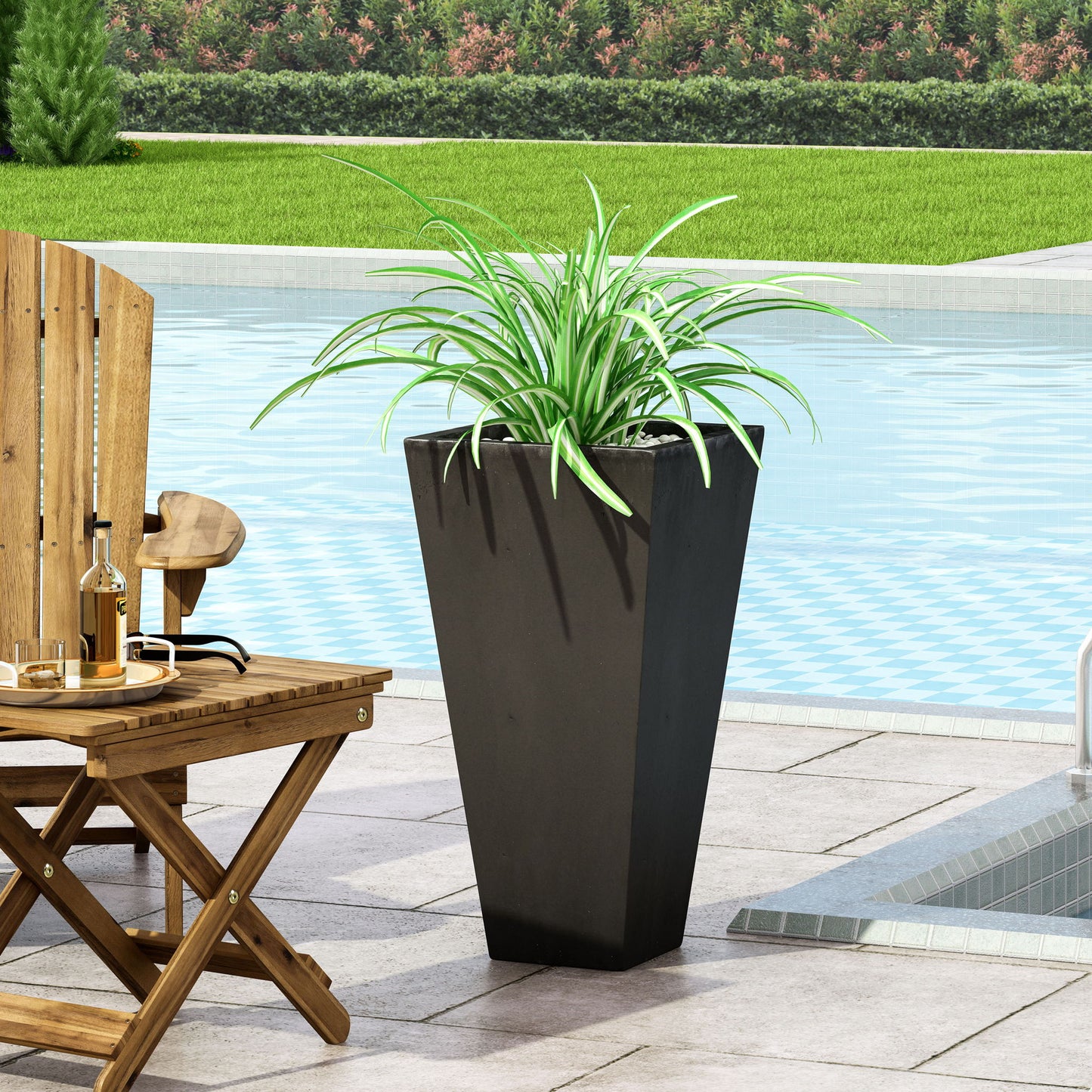 A tall, black Outdoor Modern Cast Stone Planter with a green spider plant sits by the pool on a tiled patio near a wooden table and chair, with grass and bushes in the background.