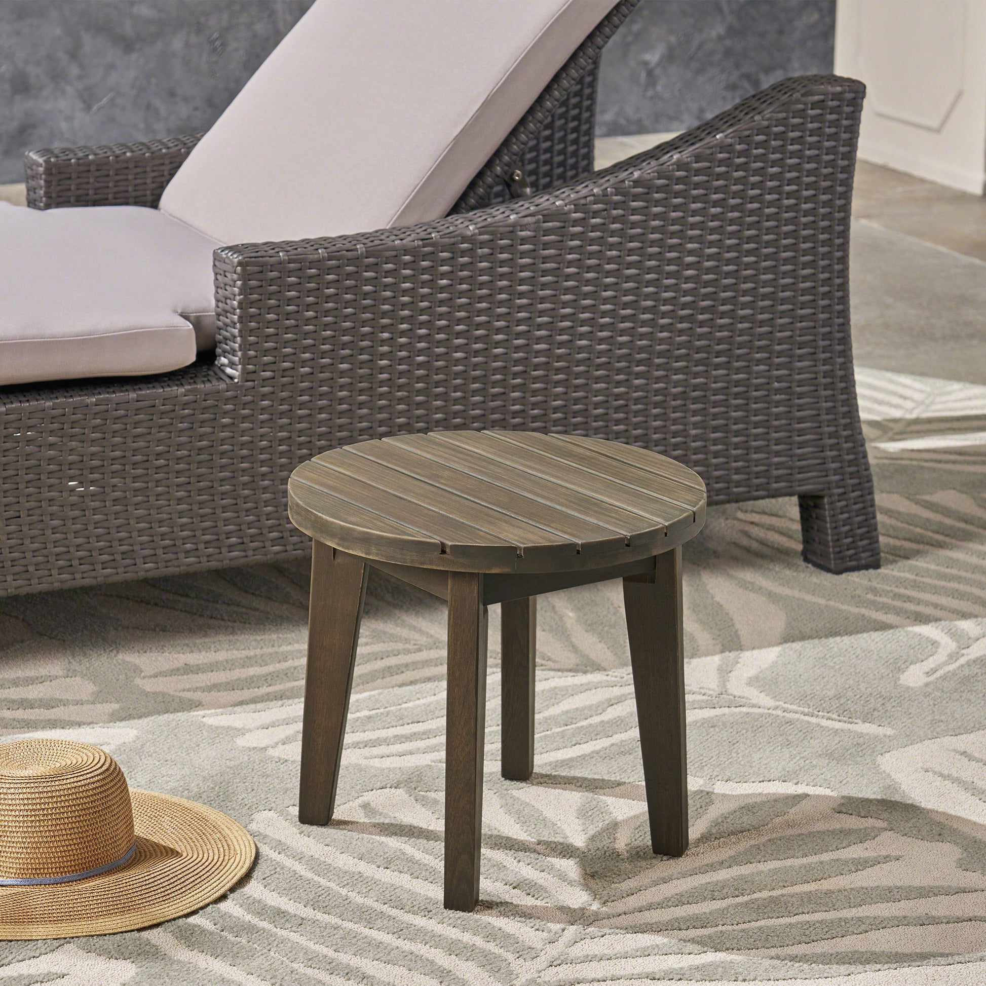 An Outdoor Acacia Wood Side Table With Weather Resistance stands on a patterned outdoor rug beside a wicker lounge chair with cushion; a straw hat rests on the ground nearby.