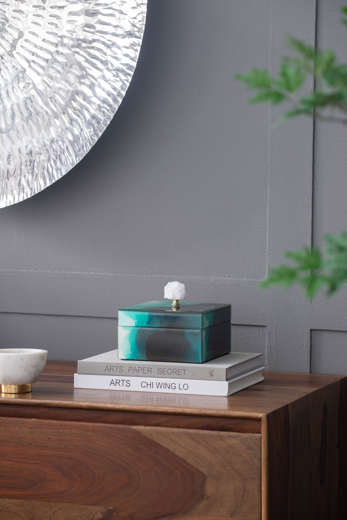 The Bethany - Aqua Jewelry Box sits on two stacked books atop a wooden cabinet next to a small white bowl, with round metallic wall art and a green plant in the background.