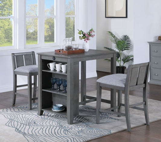 The Graham 3 Piece Small Space Counter Height Dining Table With Shelves and 2 Chairs stands on a patterned rug, displaying mugs and glasses on its shelves and a tray with a teapot and cups on top. Large windows and plants are in the background.