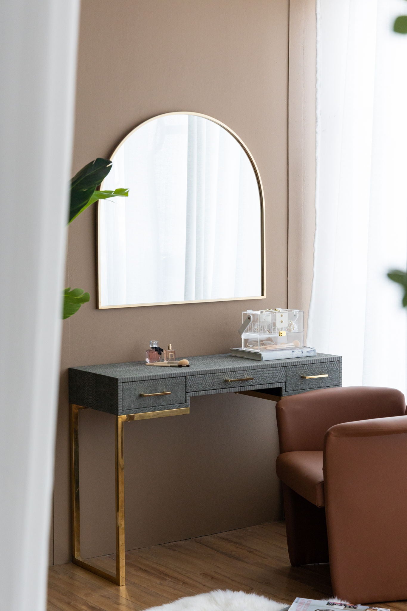 A modern vanity with a gray textured surface and gold legs sits against a beige wall. On top is the Arched Mirror With Metal Frame, along with makeup and perfume. A brown chair and sheer white curtains are nearby.