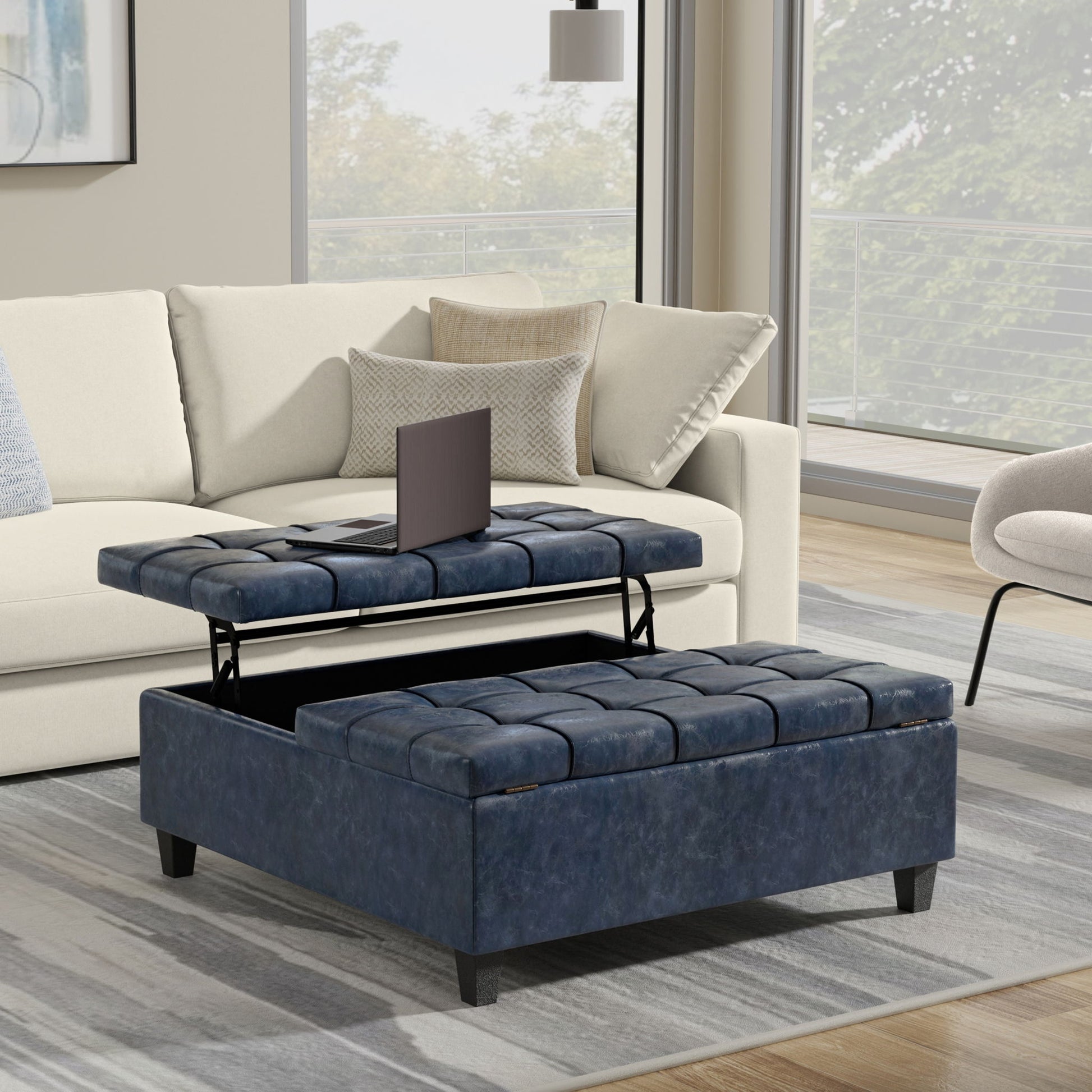 The Harrison Upholstered Coffee Table Storage Ottoman in blue, featuring a lift-top for storage, sits in front of a beige sofa in a modern living room with large windows and neutral decor.