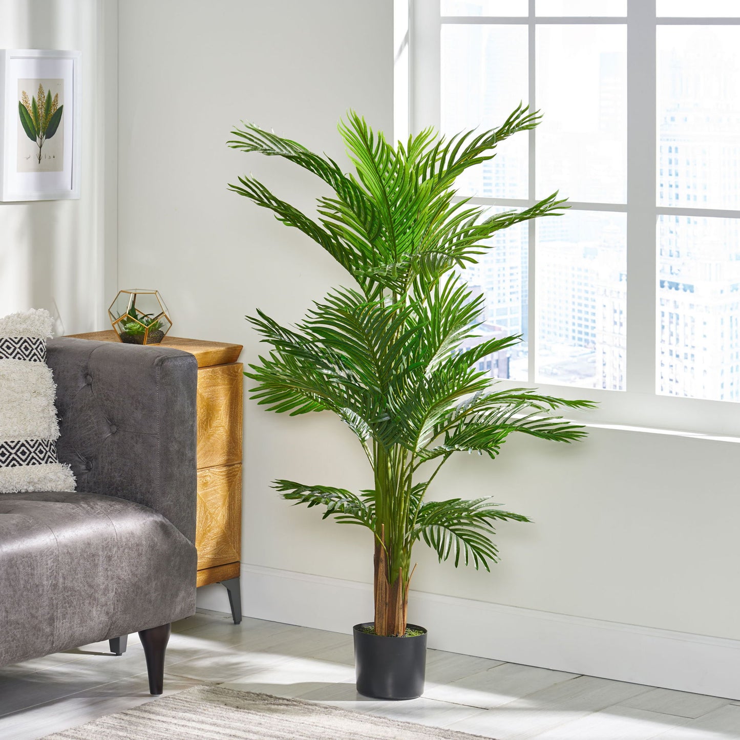 An Artificial Palm Tree in a black pot stands by a gray sofa in a bright, modern room with large windows, light walls, a small wooden dresser, and decorative accents.