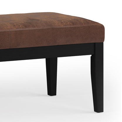 A close-up view of the Lacey - Upholstered Tufted Ottoman Bench featuring a brown cushioned seat and black wooden legs, set against a white background.