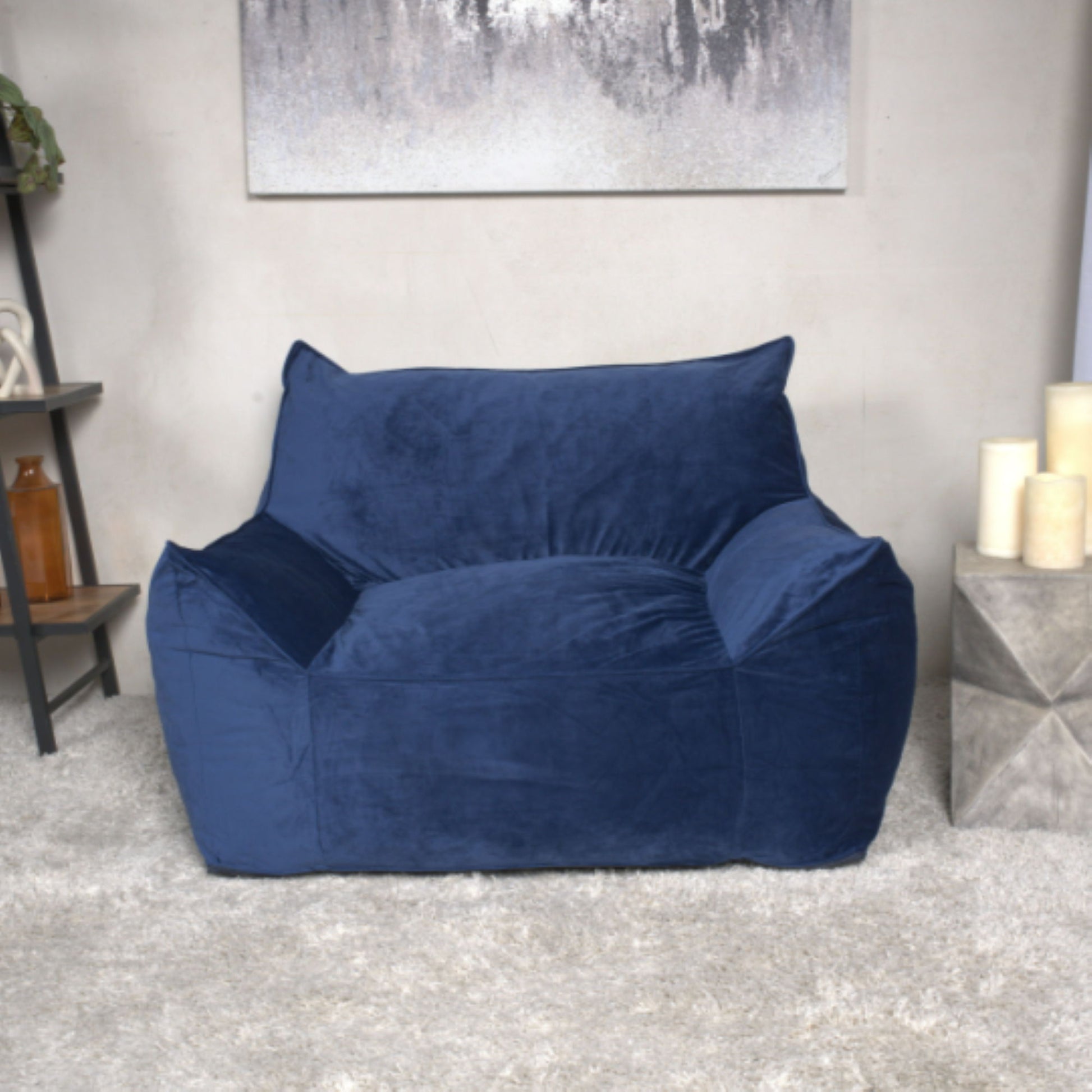 The Allea Bean Bag Chair With Armrests in deep blue sits on a light gray plush carpet, surrounded by neutral-toned decor, candles, and a shelf in the background.