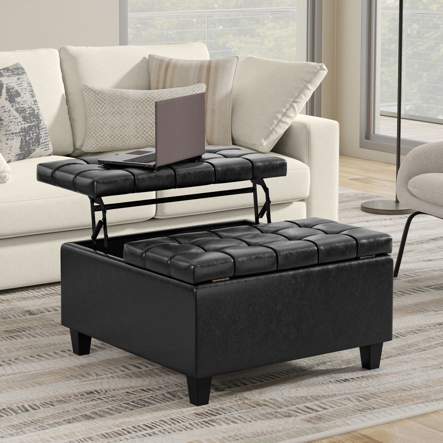 The Harrison Large Coffee Table Storage Ottoman, featuring a black tufted design and lift-top tray with a laptop, rests on a striped rug before a beige sofa with patterned cushions in a modern living room.