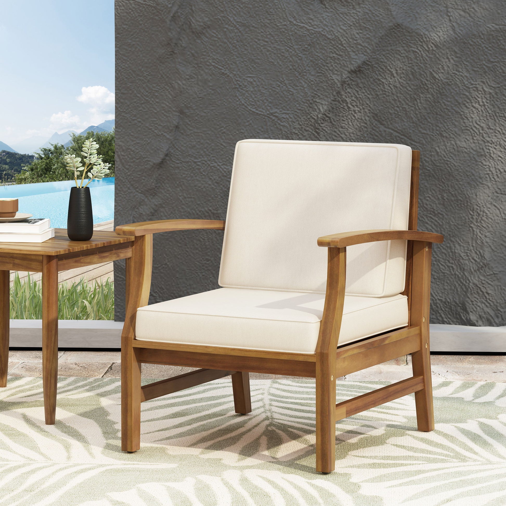 The Perla Contemporary Club Chair with water-resistant cream cushions sits on a leaf-patterned rug next to a small wooden table and black vase, set outdoors by the pool with mountains in the background.