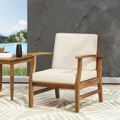 The Perla Contemporary Club Chair with water-resistant cream cushions sits on a leaf-patterned rug next to a small wooden table and black vase, set outdoors by the pool with mountains in the background.