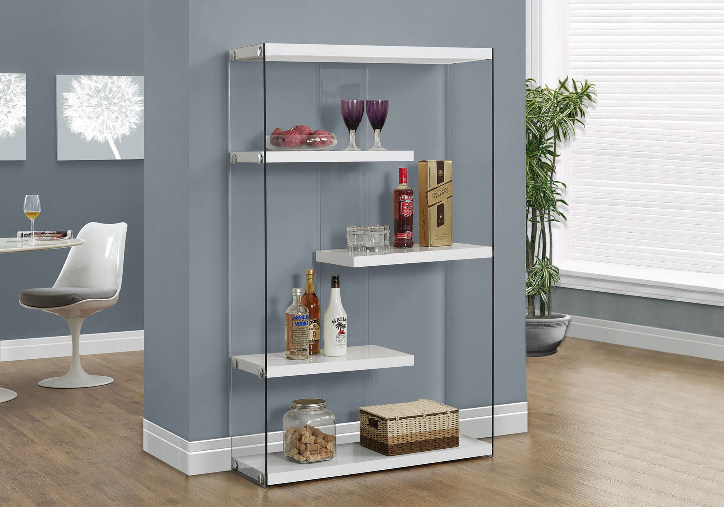 A glossy white 5-tier bookshelf displays liquor bottles, two glasses, a fruit bowl, a basket, and a jar of corks against a gray wall in a modern room with wood flooring.