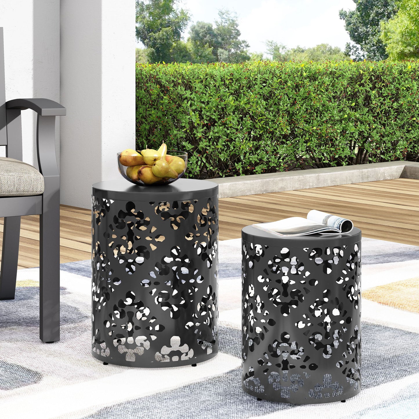 Two black Outdoor Metal Modern Design End Tables with patterned tops are on a patio—one holds a bowl of pears—next to a cushioned chair and rolled magazine, set against a green hedge and wooden deck.