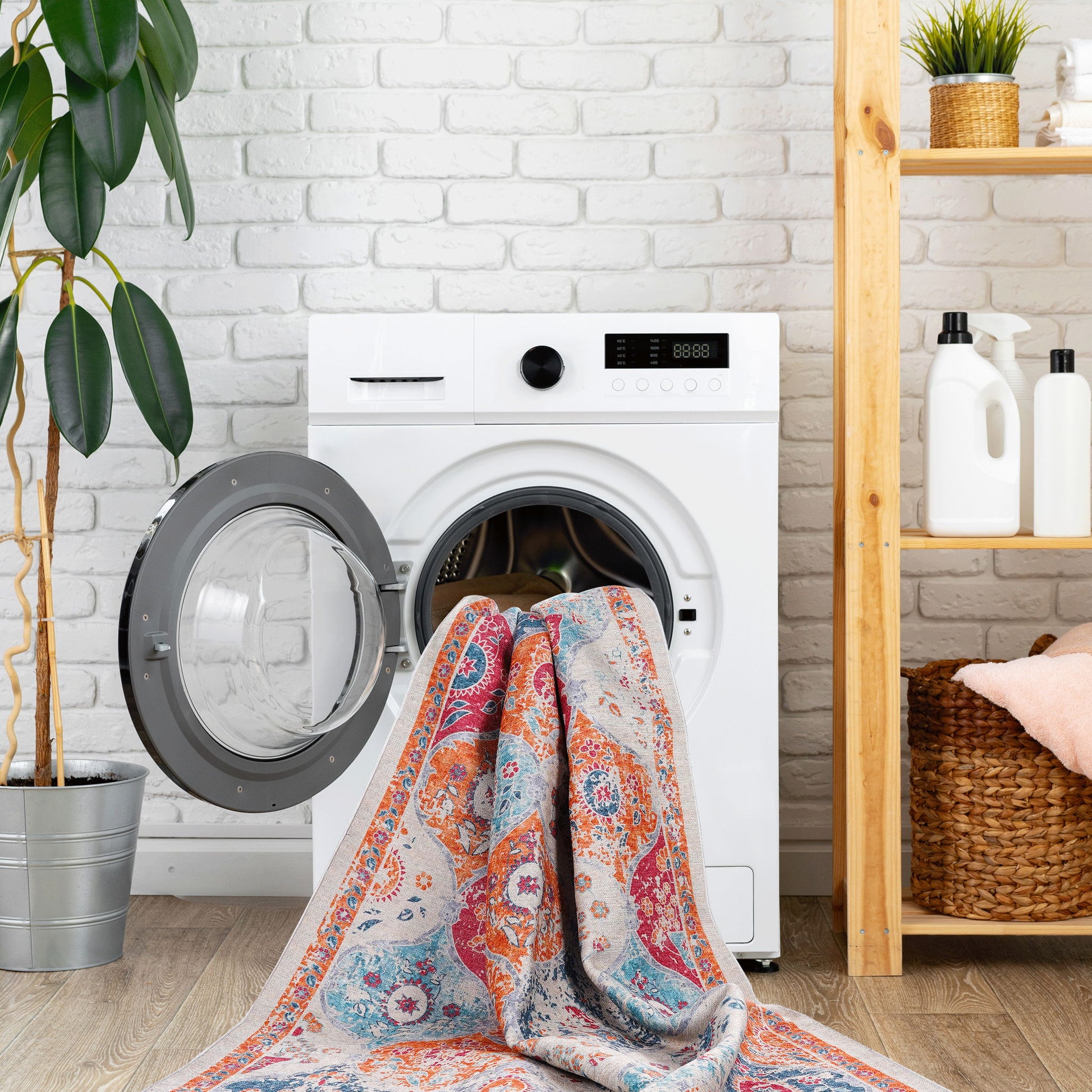 The Maval - Area Rug, featuring a colorful pattern, is partially inside a front-loading washing machine with its door open. The room also has houseplants, laundry shelves, and a white brick wall in the background.