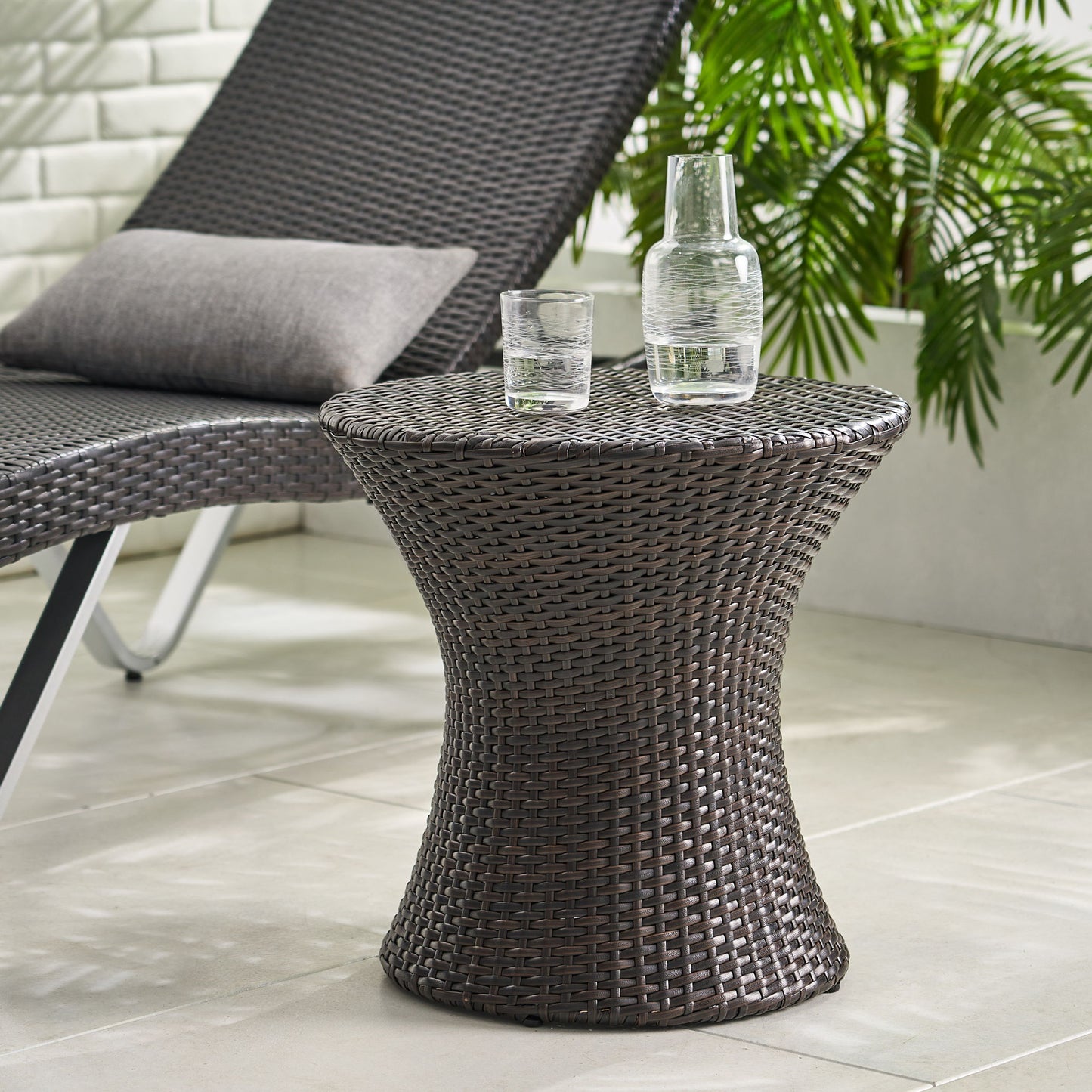 The Thrive Hourglass Rattan Table (no glass top) is placed next to a matching wicker lounge chair with a gray cushion on a tiled floor, surrounded by green plants.