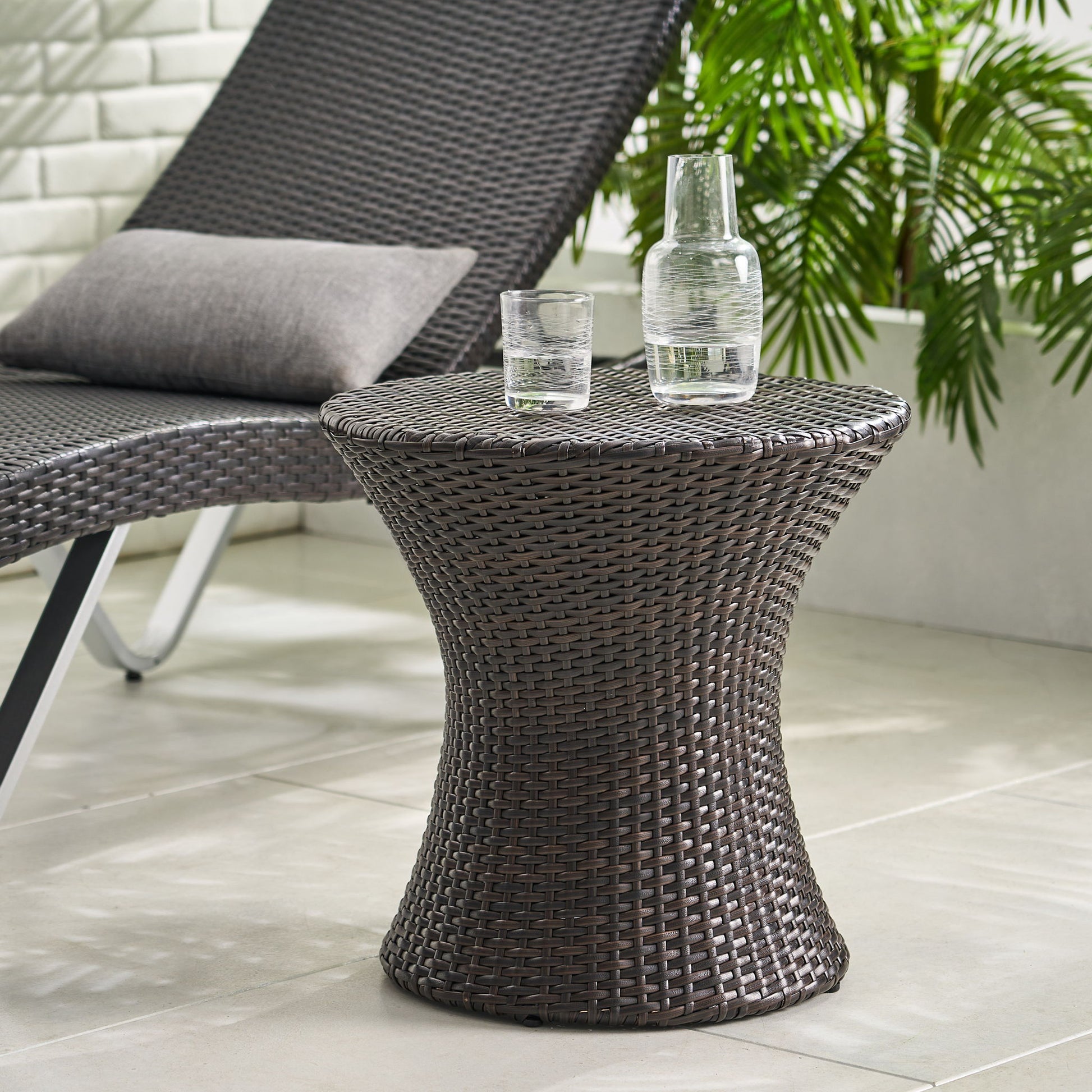 The Thrive Hourglass Rattan Table (no glass top) is placed next to a matching wicker lounge chair with a gray cushion on a tiled floor, surrounded by green plants.