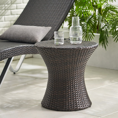 The Thrive Hourglass Rattan Table (no glass top) is placed next to a matching wicker lounge chair with a gray cushion on a tiled floor, surrounded by green plants.