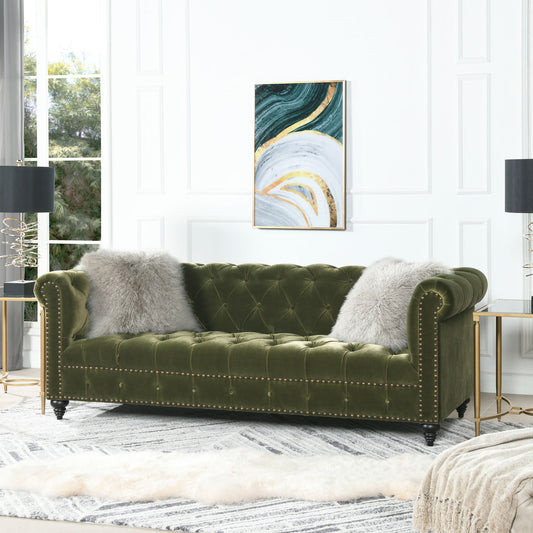 The Alto Tufted Chesterfield Sofa in green velvet with gold nailhead trim sits against a white paneled wall, adorned with two gray pillows. Abstract art hangs above, while black lamps flank the sofa over a modern rug.