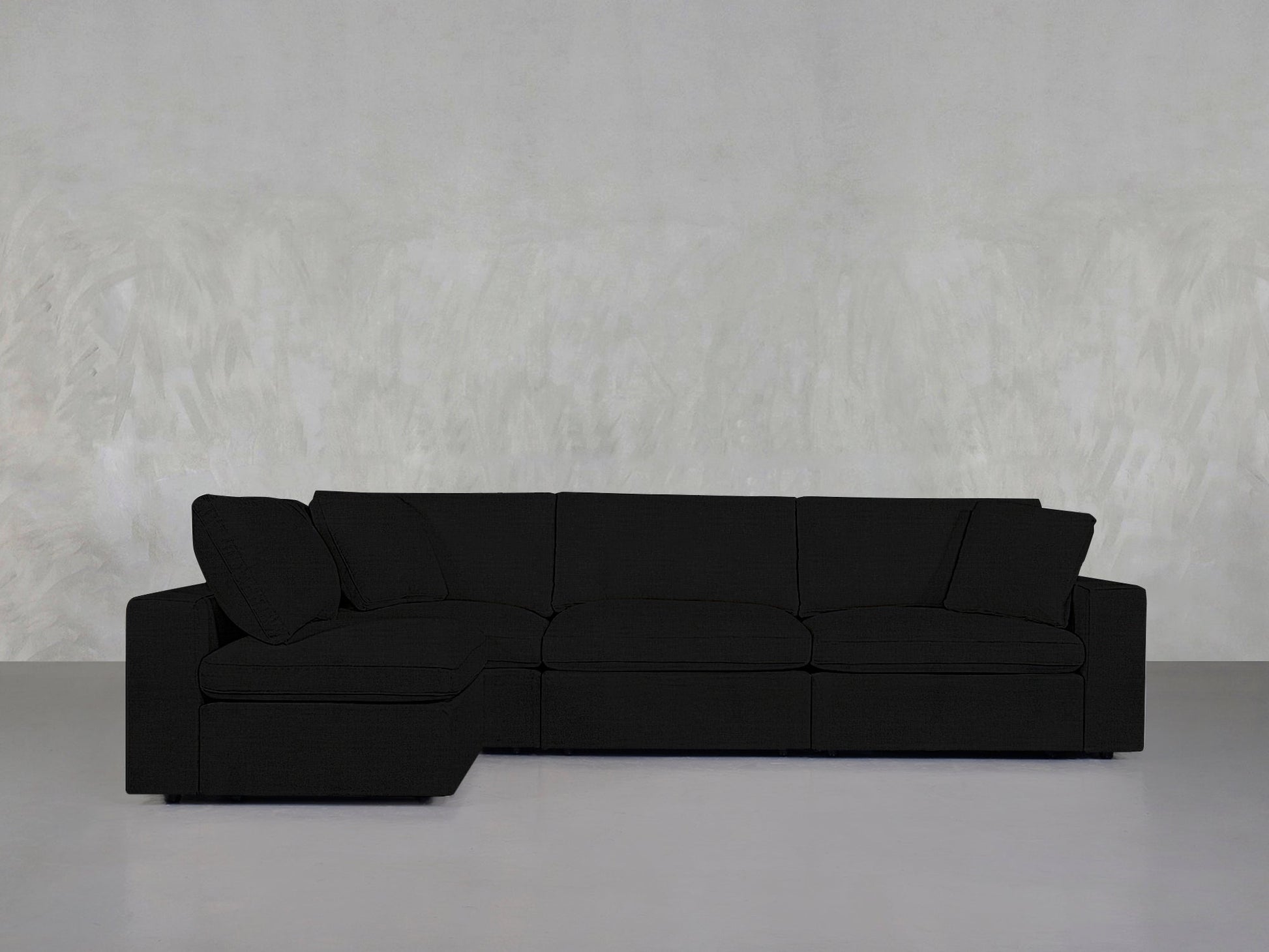 A 4-Seat Modular Open Corner Sectional with a left-facing chaise sits against a light gray textured wall and smooth gray floor in a minimalist room.