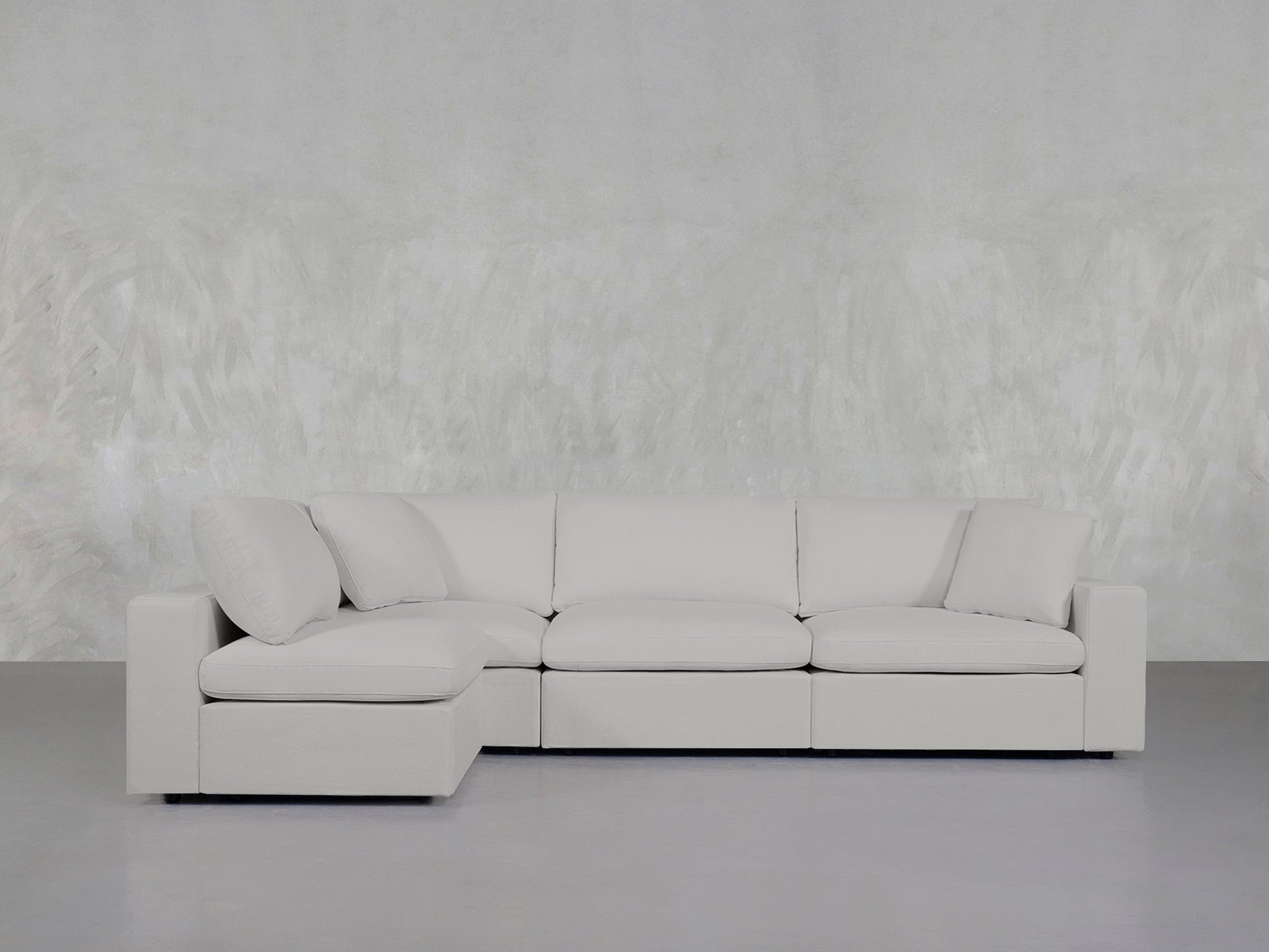 A 4-Seat Modular Open Corner Sectional in light gray, featuring clean lines and soft cushions, is set against a textured pale gray wall in a minimalist room with a smooth gray floor.