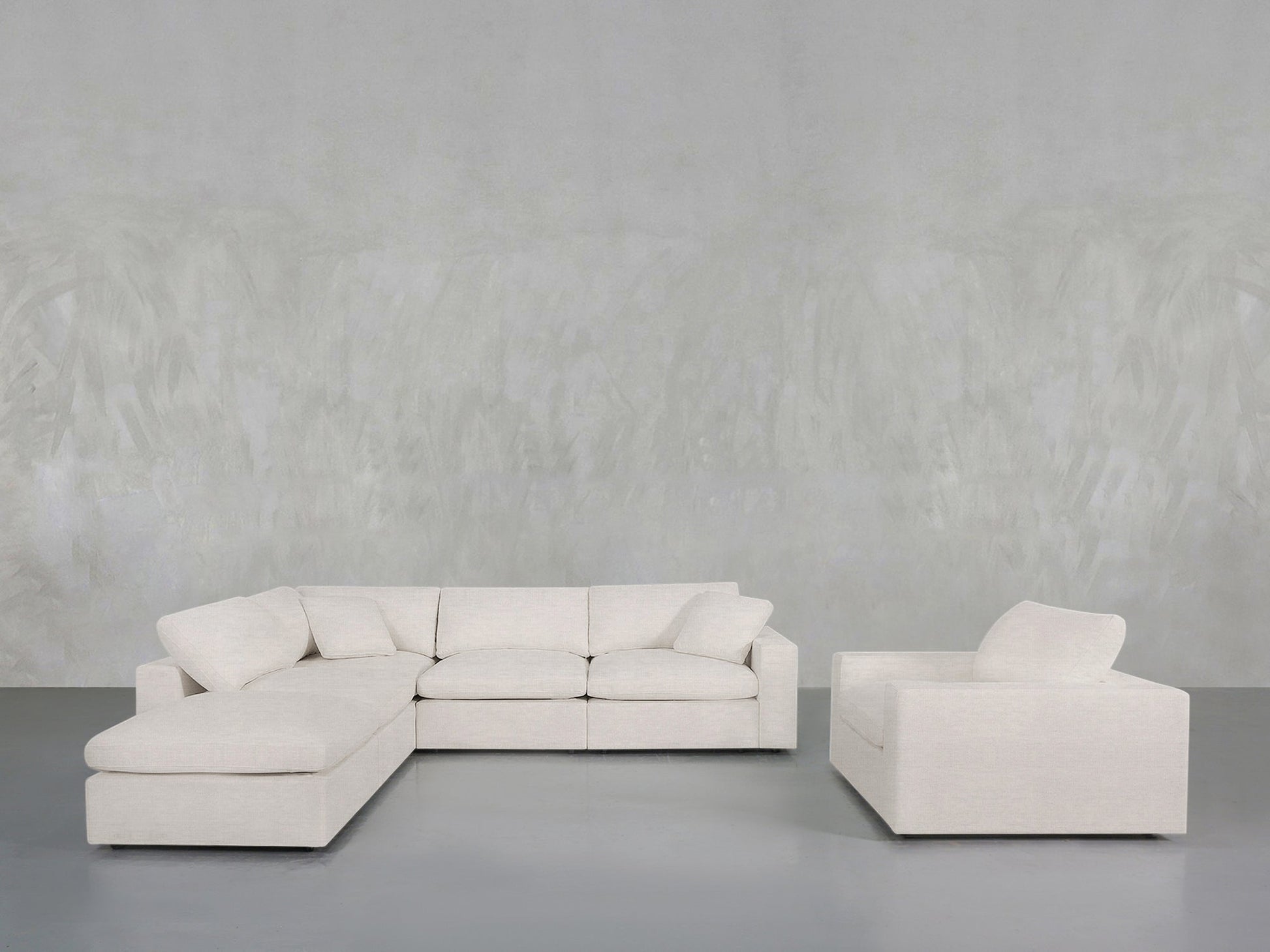 The 5-1 Sectional & Armchair Set, featuring a modern light beige sofa with plush cushions and ottoman, sits beside the matching armchair in a minimalist room with a textured gray wall and smooth gray floor.