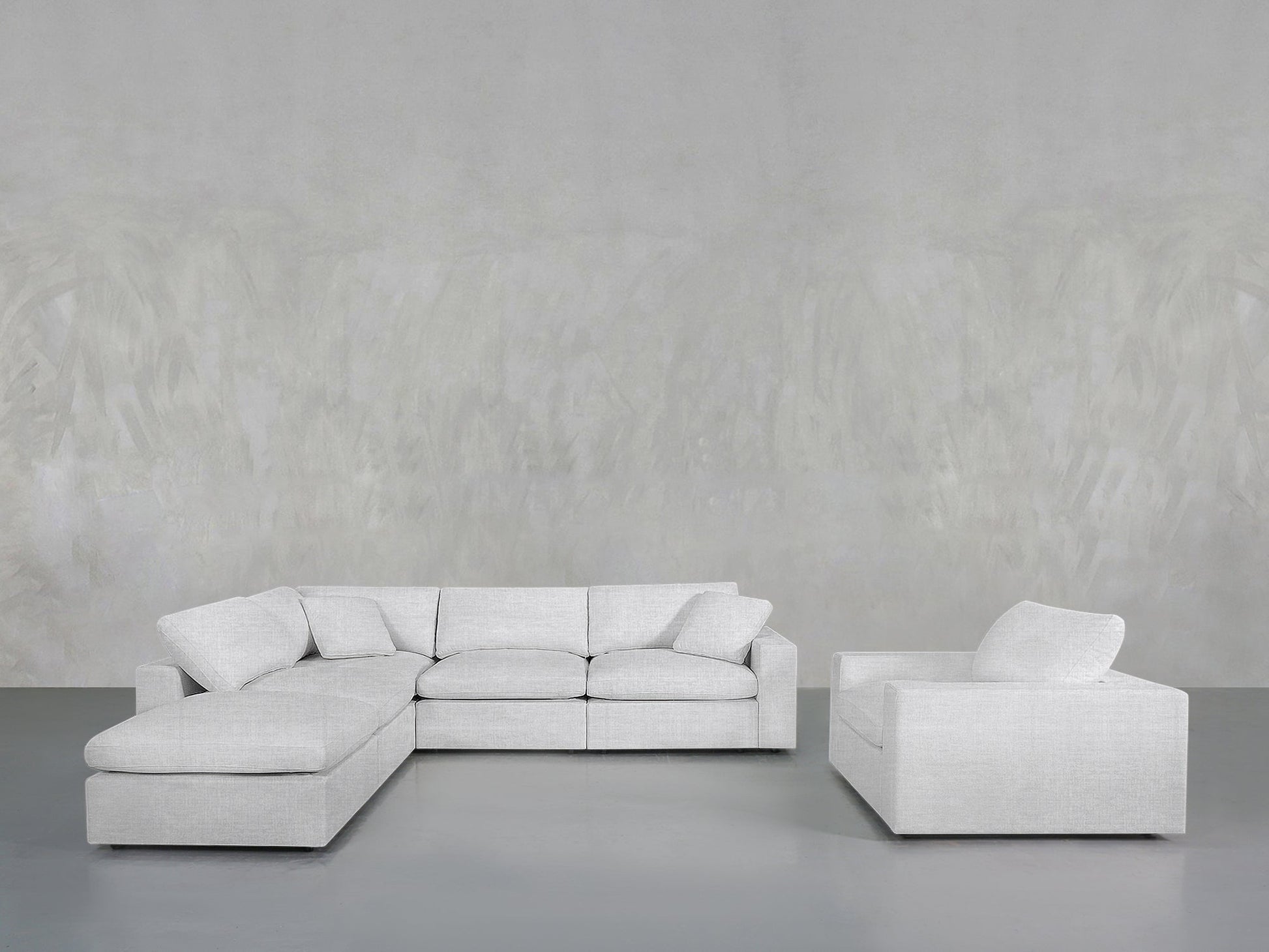 A modern living room features the 5-1 Sectional & Armchair Set in light gray, both with cushions, placed against a plain textured light gray wall and a smooth gray floor.