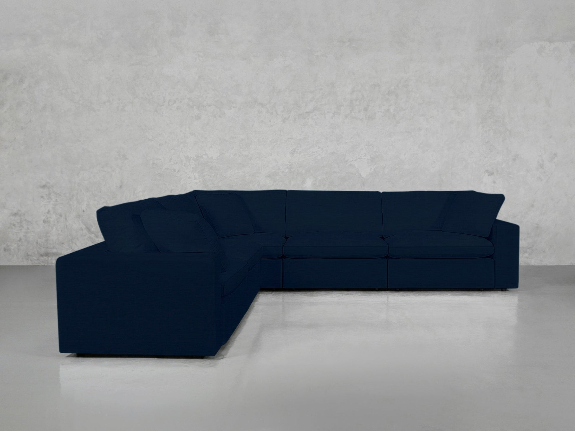A 5-Seat Modular Corner Sectional in dark blue, featuring clean lines and plush cushions, forms a right-angle L-shape in an empty room with light gray concrete walls and a polished gray floor.
