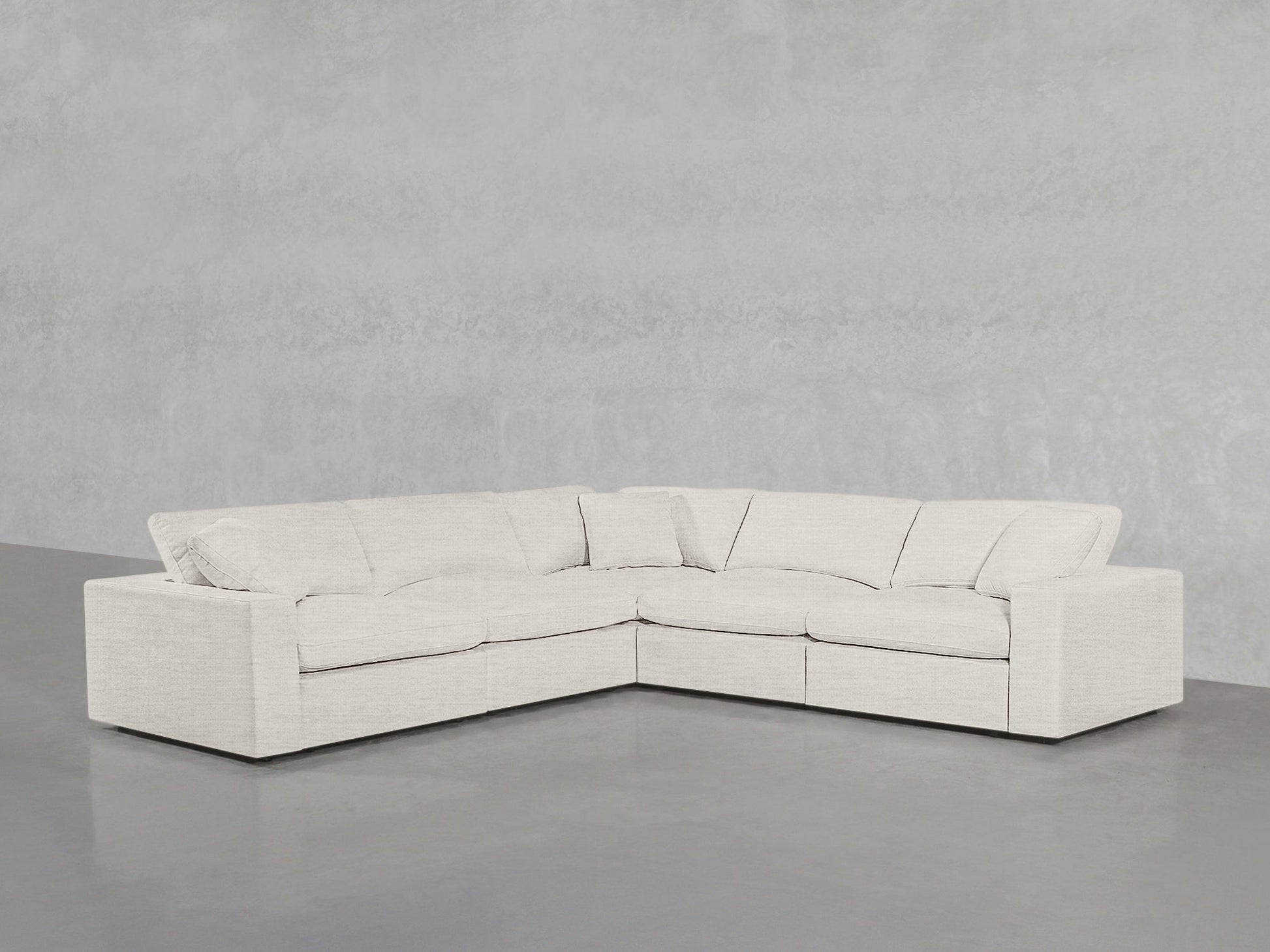 A modern, light beige 5-Seat Modular Corner Sectional with plush cushions is arranged against a plain, light gray concrete wall and floor.
