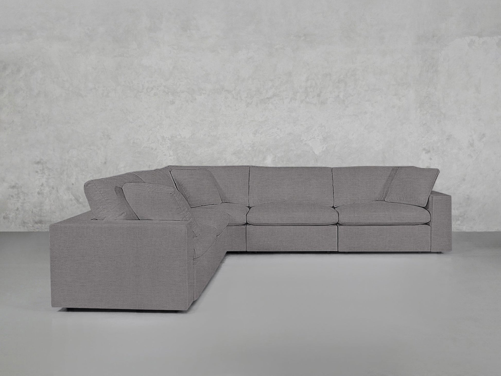 A contemporary 5-Seat Modular Corner Sectional in light gray, featuring three seat and three back cushions, positioned before a plain, light gray textured wall and floor.
