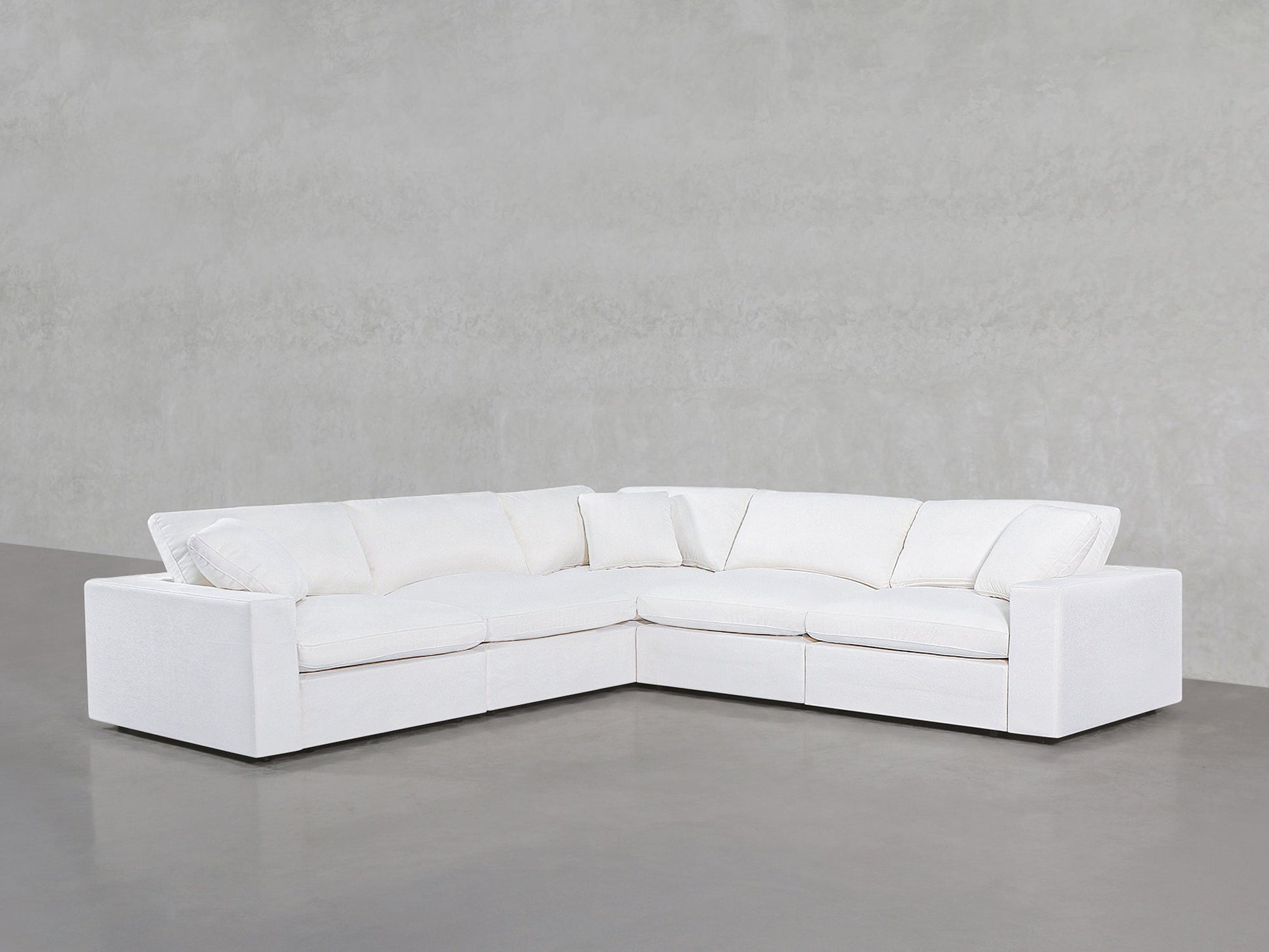 A 5-Seat Modular Corner Sectional with plush cushions is arranged in an L-shape against a light gray textured wall and gray floor in a minimalistic room.