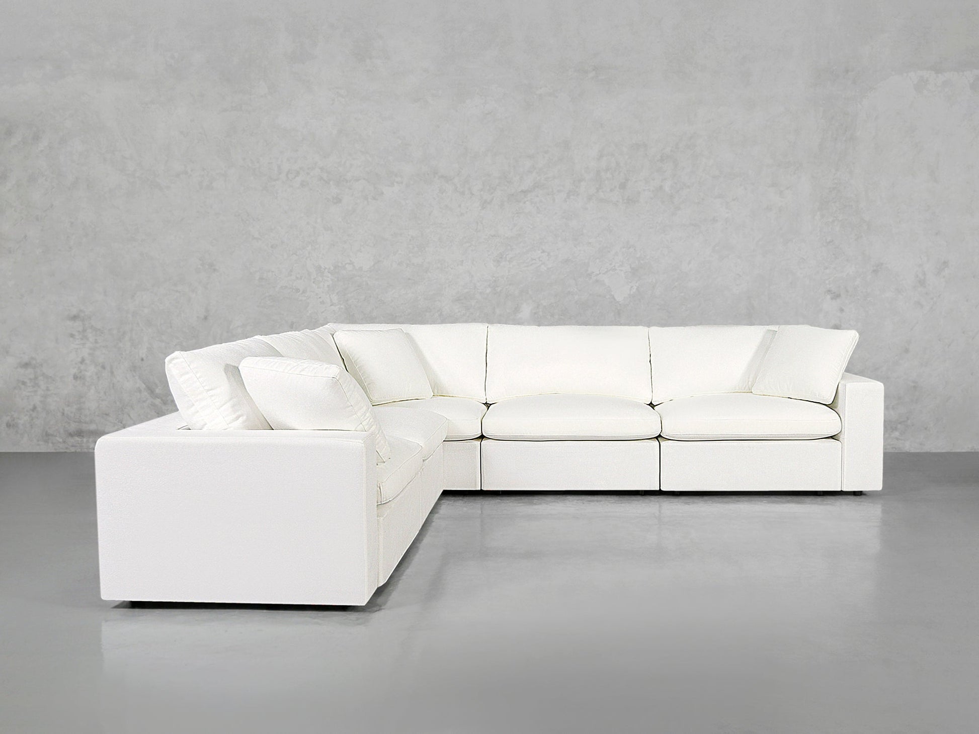 A 5-Seat Modular Corner Sectional with plush cushions sits against a plain light gray concrete wall and gray floor, creating a minimalist setting.
