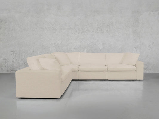 A 5-Seat Modular Corner Sectional with clean lines and plush cushions, in beige, stands against a light gray textured wall on a smooth gray floor.