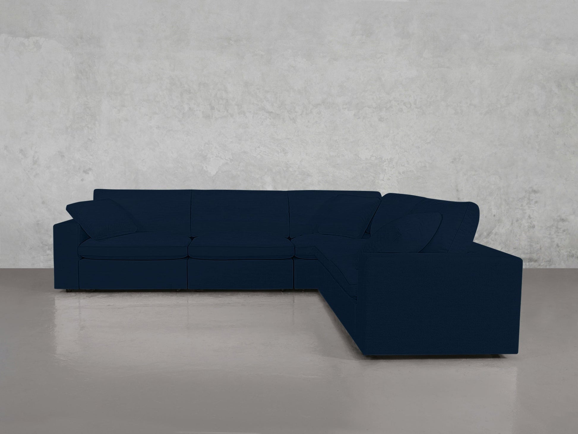 A 5-Seat Modular Corner Sectional with cushions and armrests in dark blue sits on a polished concrete floor against a light gray textured wall.