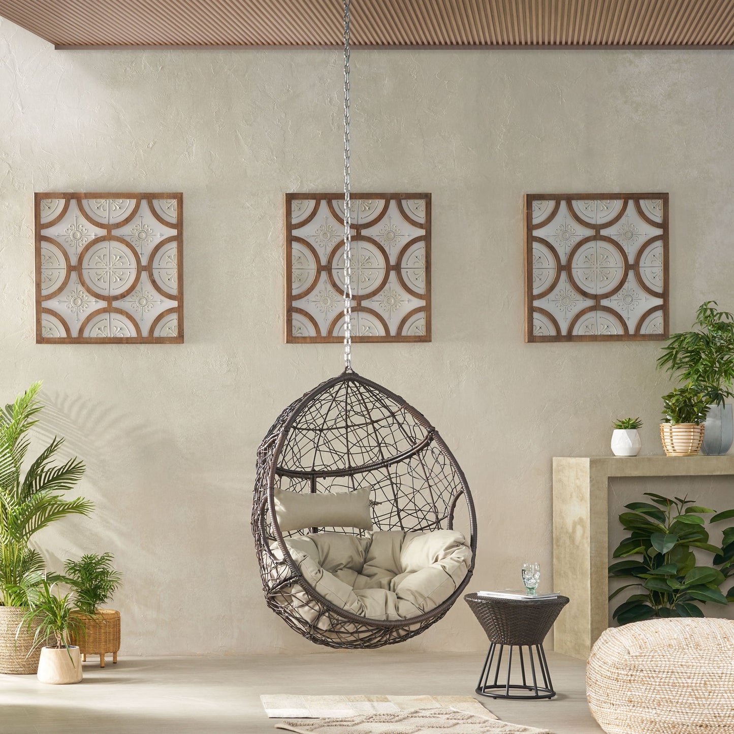 The Teardrop Hanging Chair With 8Ft Chain, featuring beige cushions, is suspended in a modern living room with geometric framed art, lush plants, a small side table, and a light-colored floor.