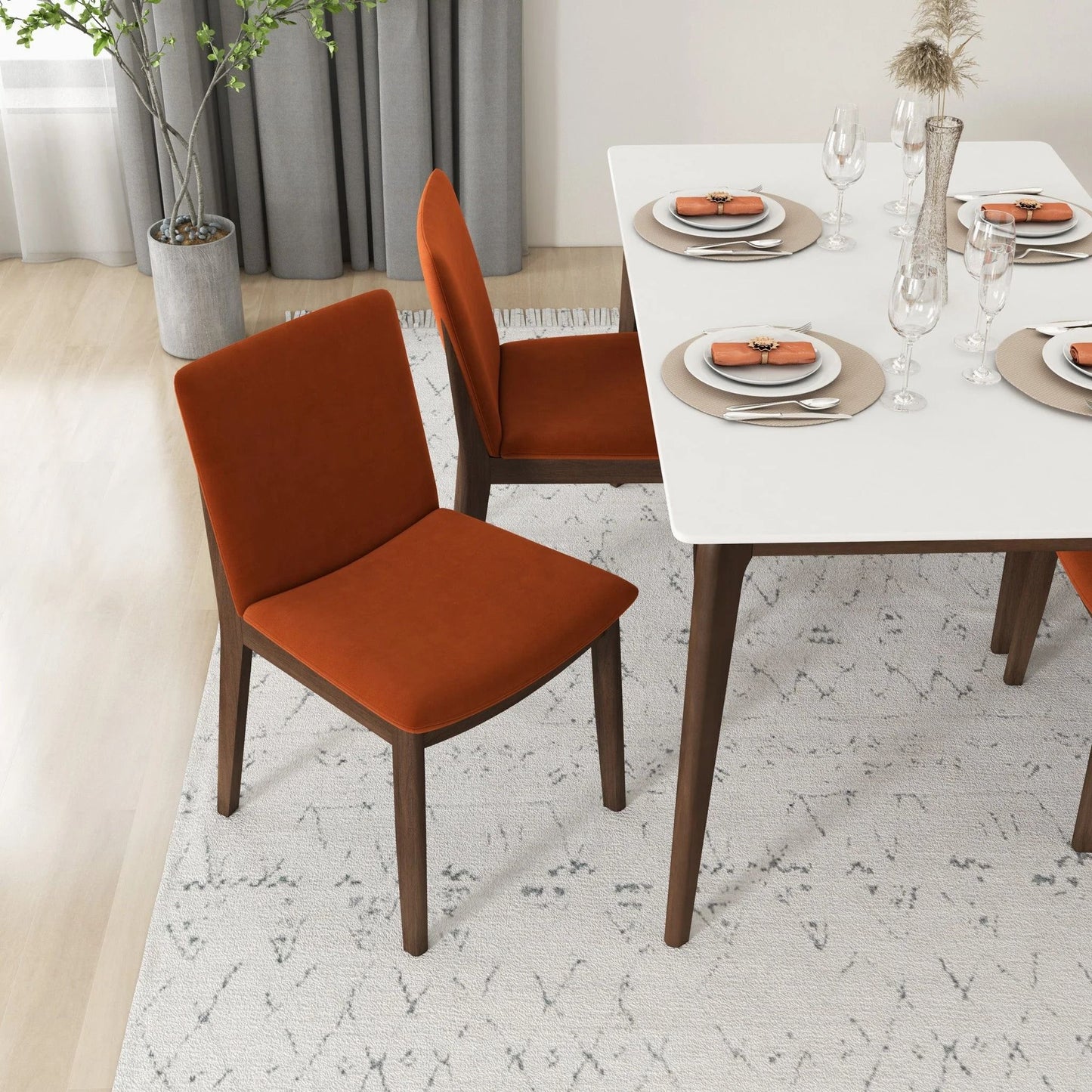 A modern dining room with a white table for four, featuring Laura - Mid-Century Modern Solid Wood Dining Chairs in orange, neatly set plates and glassware, and a potted plant in the corner on a light patterned rug.