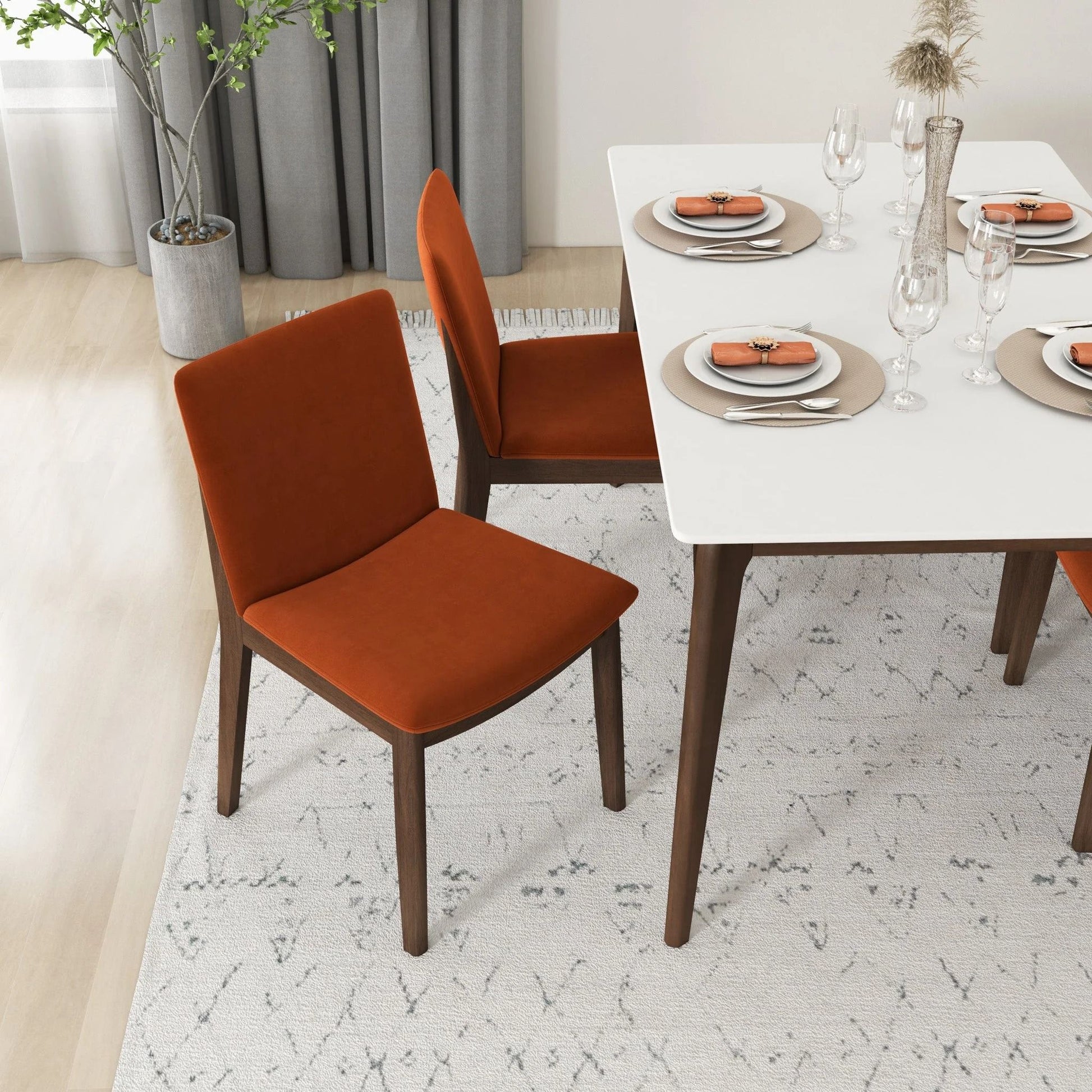 A modern dining room with a white table for four, featuring Laura - Mid-Century Modern Solid Wood Dining Chairs in orange, neatly set plates and glassware, and a potted plant in the corner on a light patterned rug.