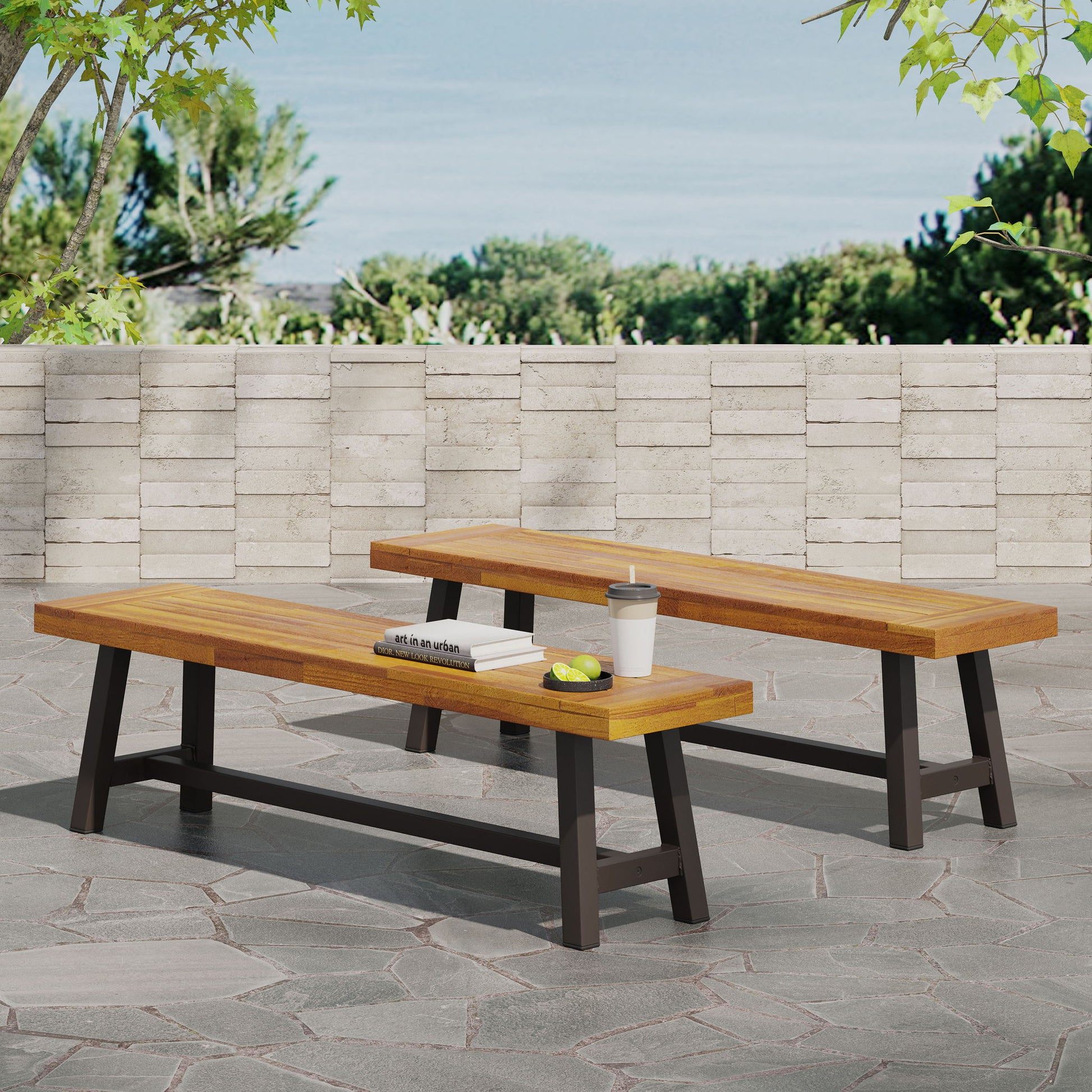 Two Tranquilizer Outdoor Acacia Wood Benches with slat panel design and black legs sit on a stone patio, one topped with books, an apple, a tennis ball, and a travel cup. A stone wall and trees appear in the background beneath a blue sky.