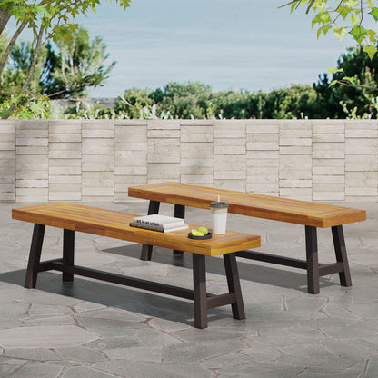 Two Tranquilizer Outdoor Acacia Wood Benches with slat panel design and black legs sit on a stone patio, one topped with books, an apple, a tennis ball, and a travel cup. A stone wall and trees appear in the background beneath a blue sky.