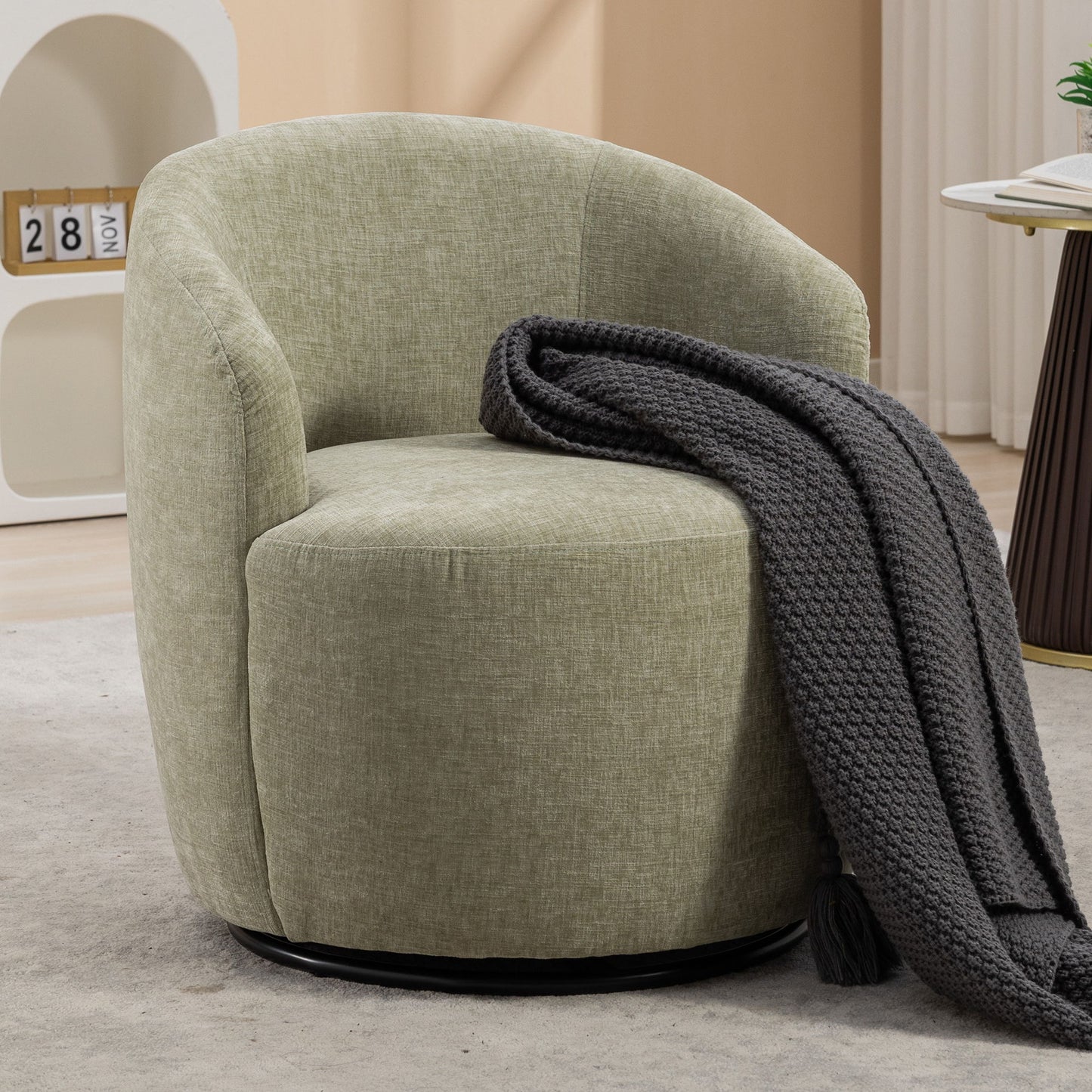 A light green Chenille Fabric Swivel Accent Armchair Barrel Chair with a curved backrest and powder-coated metal ring sits on a light carpet. A dark gray knitted throw blanket is draped over one side, with a small shelf and table in the background.