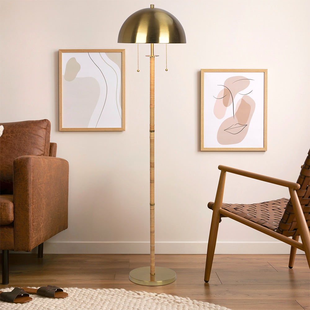 A modern living room featuring a brown leather sofa, woven chair, abstract wall art, wooden flooring, sandals on a cream rug, and the Allure 2-Light Floor Lamp in gold with natural rattan tube and double pull chain.