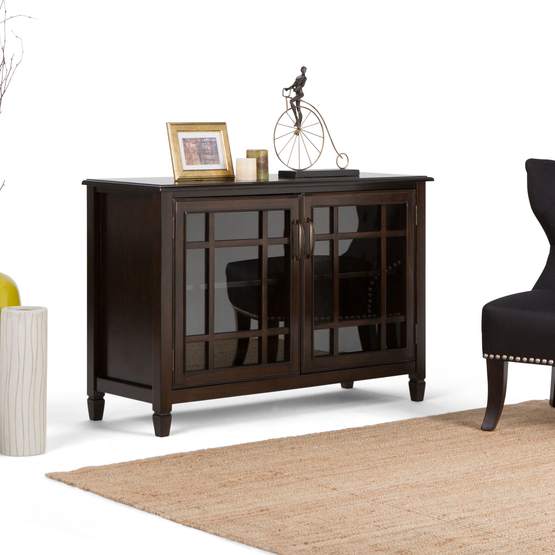 The Connaught - Handcrafted Low Storage Cabinet with glass-paneled doors stands on a beige rug beside a black chair, topped with framed photos, candles, and a decorative metal bicycle sculpture.