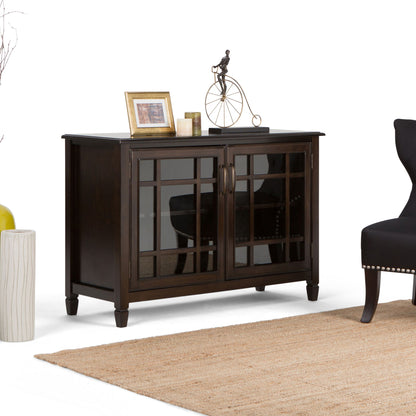 The Connaught - Handcrafted Low Storage Cabinet with glass-paneled doors stands on a beige rug beside a black chair, topped with framed photos, candles, and a decorative metal bicycle sculpture.