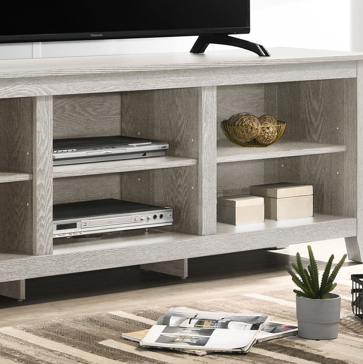 The Benito TV Stand With Open Shelves And Cable Management in light gray displays electronics, gold spheres, small boxes, and features a nearby potted cactus. A partial TV and striped rug are also in view.