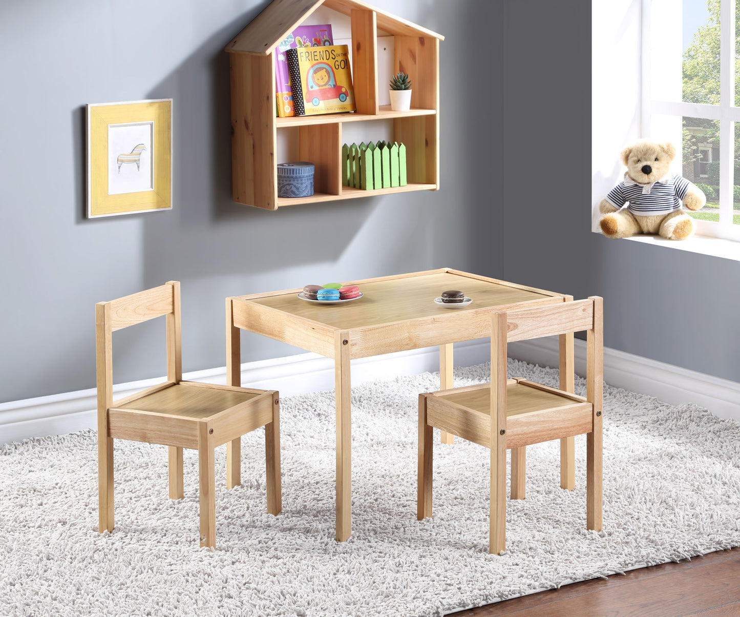 The Della 3 Piece Solid Wood Kids Table & Two Chair Set in natural finish sits on a white rug in a bright room. A teddy bear is on the window sill, books and toys rest on a wall shelf, and framed art decorates the gray wall.
