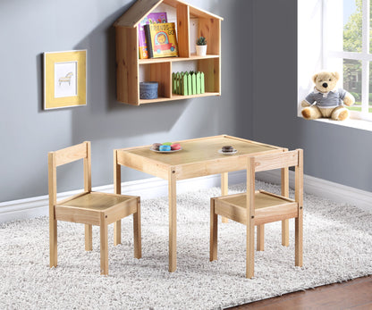 The Della 3 Piece Solid Wood Kids Table & Two Chair Set in natural finish sits on a white rug in a bright room. A teddy bear is on the window sill, books and toys rest on a wall shelf, and framed art decorates the gray wall.