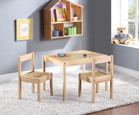 The Della 3 Piece Solid Wood Kids Table & Two Chair Set in natural finish sits on a white rug in a bright room. A teddy bear is on the window sill, books and toys rest on a wall shelf, and framed art decorates the gray wall.